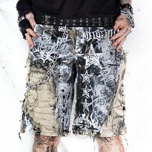 May include: Distressed black and white shorts with a star and graffiti design. The shorts have a ripped and frayed hem and are paired with a black belt. The shorts are part of a punk or alternative fashion style.