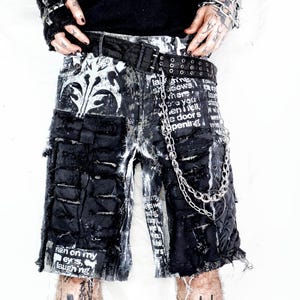 May include: Distressed black and white denim shorts with a chain. The shorts have a black belt, patches, and text. The design includes a white floral pattern and the text "rain on my eyes, laughing".