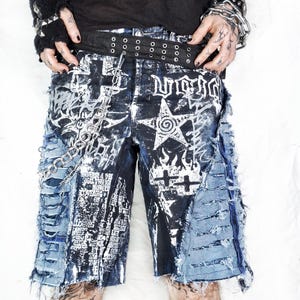 May include: Distressed denim shorts with a black and white graphic print, featuring a star and cross design. The shorts have ripped details and a chain accent. A black belt with silver grommets is visible.