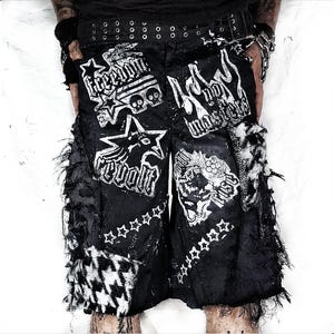 May include: Black shorts featuring white graphic prints with the text Freedom, Revolt, No Masters, and Rise. The shorts include a studded belt, frayed side panels, and black and white houndstooth fabric patches on the lower front sections.