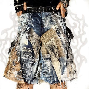 May include: Distressed denim shorts featuring a dark blue wash with heavy white paint splatters, frayed fabric patches, and layered textile textures. The garment includes metal chain attachments, a black studded belt, and printed text.
