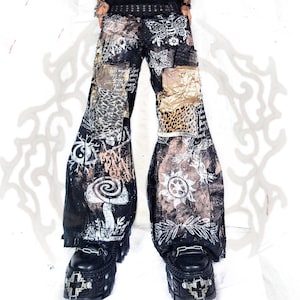 May include: Black wide-leg pants featuring a collage of white screen-printed graphics including a moth, a mushroom, a sun, and text. The pants incorporate distressed patches with leopard print and metallic gold fabric accents, paired with black platform shoes.