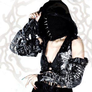 May include: A black hooded garment featuring distressed fabric with white graphic prints and silver-toned spikes along the hood edge. The piece includes long sleeves with frayed edges and a cropped bodice design, showcasing a dark, textured aesthetic.