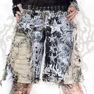 Puede incluir: Pantalones cortos desgastados en beige y negro con una cadena de metal pesada sujeta a un cinturón con ojales. La tela muestra un estampado gráfico de alto contraste con estrellas blancas, letras tipo grafiti y paneles deshilachados.