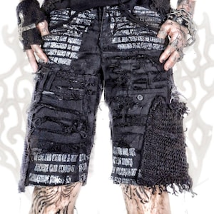May include: Black distressed shorts featuring a white printed text pattern across the fabric. The garment includes multiple frayed horizontal panels, a single button closure, and a textured, dark mesh-like pocket overlay on the left leg.