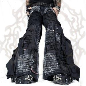 Könnte beinhalten: Schwarze Hose mit weitem Bein, distressed Stoffeinsätzen, ausgefransten Kanten und silberfarbenen Metallketten an der Taille. Das Kleidungsstück verfügt über mehrere Cargotaschen, geschichtete Stoffstreifen und weiße Textdruckmuster.