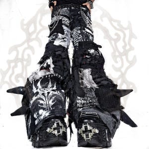 Puede incluir: Pantalones negros y blancos con aspecto desgastado, diseño de parches con texto impreso, bordes deshilachados y detalles de cadenas metálicas. Los pantalones se combinan con botas de plataforma negras con herrajes en forma de cruz plateada.