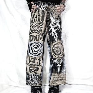 May include: Distressed denim pants with various black and white graphics, including stars, spirals, and text. The pants feature a wide leg design and are paired with black platform shoes. The word "IMMORTAL" is visible on the left leg.
