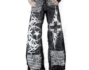 May include: Black denim pants with white tribal and cross designs. The pants have a distressed look with a belt and a wide leg. The design includes a star and text details. The pants are worn with black shoes.