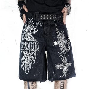 May include: Black denim shorts with white graphic designs and the word "VOID" printed on the front. The shorts feature a black belt with silver grommets and a silver chain. The shorts have a distressed look.