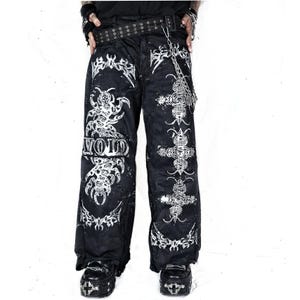 Peut inclure: Pantalon noir à jambes larges avec des motifs graphiques blancs représentant des insectes et des symboles en forme de croix. Le pantalon comprend une ceinture noire à œillets métalliques et une chaîne argentée. Le mot VOID est imprimé en blanc.