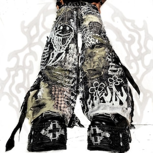 May include: Wide-leg pants featuring a distressed patchwork design in black, white, and olive green. The fabric includes printed graphics of a smiley face, flowers, flames, and handwritten text. The pants are paired with black platform shoes.