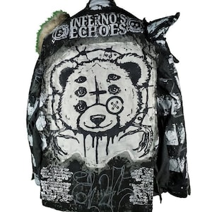 Pode incluir: Casaco preto e branco com uma estampa gráfica de um urso com vários olhos e uma cruz no peito. O casaco tem o texto "Inferno's Echoes" nas costas.
