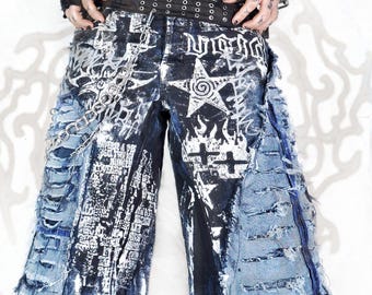 May include: Distressed denim shorts in dark blue and black featuring white graphic prints of stars, crosses, and text. The shorts include heavy fraying, side panels with horizontal slits, a black grommet belt, and a silver metal chain attachment.