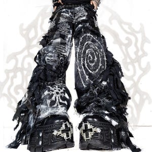 Könnte beinhalten: Schwarz-weiße, distressed Hose mit weitem Bein, ausgefransten Stoffstreifen, aufgedrucktem Text und einer großen Spiralgrafik. Die Hose wird mit schwarzen Plateaustiefeln kombiniert, die über kreuzförmige Metallbeschläge und silberne Nieten verfügen.