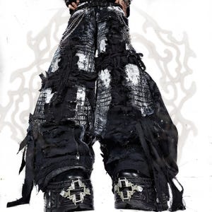 Könnte beinhalten: Schwarz-weiße Hose mit weitem Bein im Distressed-Look, mit stark ausgefransten Kanten, überlagerten Stoffbahnen und silberfarbenen Metallketten an der Taille. Dazu werden schwarze Plateaustiefel mit auffälligen silbernen Kreuzbeschlägen getragen.