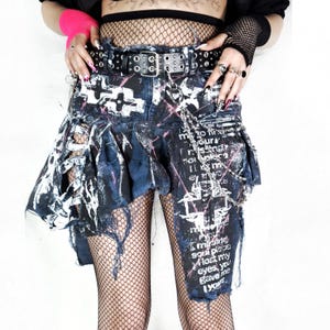 May include: Distressed denim skirt with a black belt and silver chain details. The skirt has a ripped design with white and pink paint splatters and printed text. The skirt is worn over fishnet stockings.