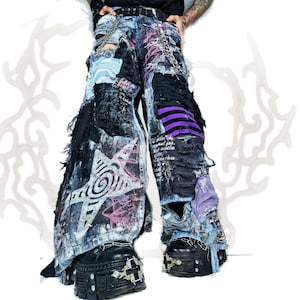 Op de afbeelding: Versleten wide-leg spijkerbroek met een grote witte stergrafiek, paarse gestreepte stoffen panelen, zwarte stoffen patches, gerafelde randen en zilverkleurige metalen kettingen aan de taille. De broek wordt gedragen met zwarte plateau-laarzen.