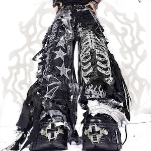 Könnte beinhalten: Schwarze Hose mit weitem Bein, abgenutztem Stoff, ausgefransten Kanten und weißen Grafikdrucken von Sternen, einem Brustkorb und Flammen. Die Hose wird mit schwarzen Plateaustiefeln kombiniert, die ein silberfarbenes, kreuzförmiges Metallemblem tragen.