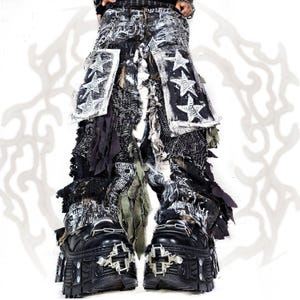 May include: Distressed black and white pants featuring frayed fabric layers, patches with white star graphics, and hanging fabric strips. The pants are paired with black platform boots that have silver-toned metal cross hardware and chain details.