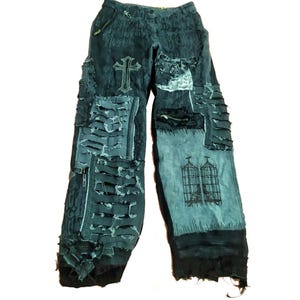 May include: Dark blue denim trousers with distressed details, featuring a cross and a cage design. The trousers have multiple layers of fabric and frayed edges.