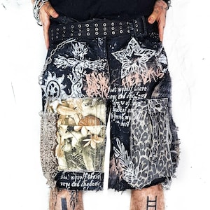 May include: Black denim shorts featuring a double-prong grommet belt, a chain attachment, and distressed edges. The fabric displays white graphic prints of stars, crosses, and text, alongside a floral patch pocket and a leopard print pocket.