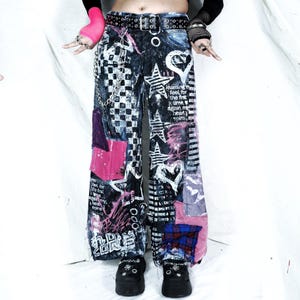May include: Dark wash denim jeans with a patchwork design. The jeans feature pink, purple, and black patches, along with white star and heart graphics. Text is printed on the jeans. The jeans have a wide leg and a black belt.