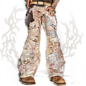 May include: Beige distressed pants featuring a wide-leg silhouette with heavy horizontal shredding and fraying. The fabric is layered with white printed text and graphics. A tan belt with a large buckle is worn at the waist with hanging brown beads.