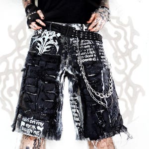 Op de afbeelding: Zwart-witte distressed shorts met een grafische print met de tekst "rain on my eyes, laughing" en "where you when I fell, the door is opening". De shorts zijn voorzien van een zwarte riem, metalen ketting en gerafelde zoomdetails.
