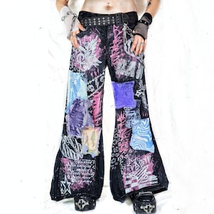 May include: Black denim jeans with a wide leg, featuring patches of various colors and textures. The jeans are embellished with graffiti-style designs in pink and white, and a black belt with silver grommets.