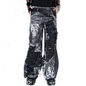 May include: Black distressed denim trousers with white graphic designs and text. The trousers feature a wide leg, multiple rips, and a studded belt. The design includes star patterns and ornate floral motifs, creating a gothic aesthetic.