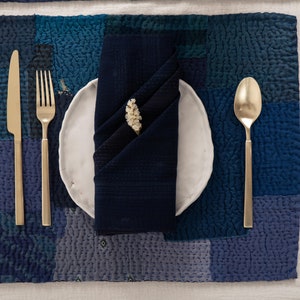 Könnte beinhalten: Ein weißer Keramikteller mit einer blauen Serviette, die darin gefaltet ist, ein goldenes Messer und eine Gabel sowie ein kleiner Zweig weißer Blumen. Der Teller liegt auf einem blauen und weißen Patchwork-Platzset.