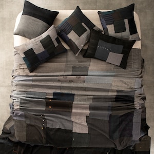 May include: A patchwork quilt in shades of grey and blue. The quilt is made up of many small squares and rectangles, and it has a rustic, handmade look. The quilt is topped with five pillows, all of which are also made of patchwork fabric.