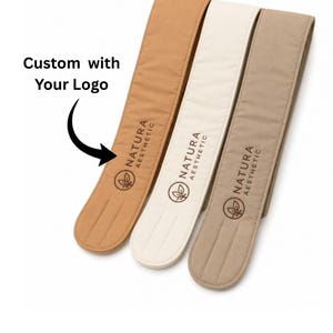 Op de afbeelding: Drie lange, rechthoekige stoffen hoofdbanden in de kleuren bruin, crème en taupe. Elke hoofdband is bedrukt met de tekst 'NATURA AESTHETIC' en een bladlogo. De tekst 'Custom with Your Logo' wijst met een pijl naar de bruine hoofdband.