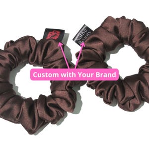 Può includere: Due scrunchies in raso marrone con un'etichetta nera che dice "Native Beauty" su uno e una stampa di labbra rosse sull'altro. Il testo "Custom with Your Brand" è scritto in rosa su sfondo bianco.