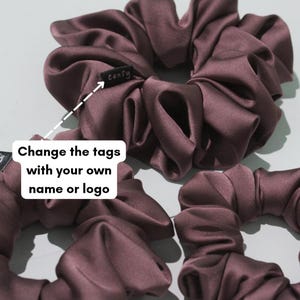 May include: Three dark plum-coloured satin hair scrunchies with gathered fabric. One scrunchie features a small black fabric tag with the white text "confy" stitched onto it. The image includes a white graphic overlay with the text "Change the tags with your own name or logo" and an arrow pointing to the tag.