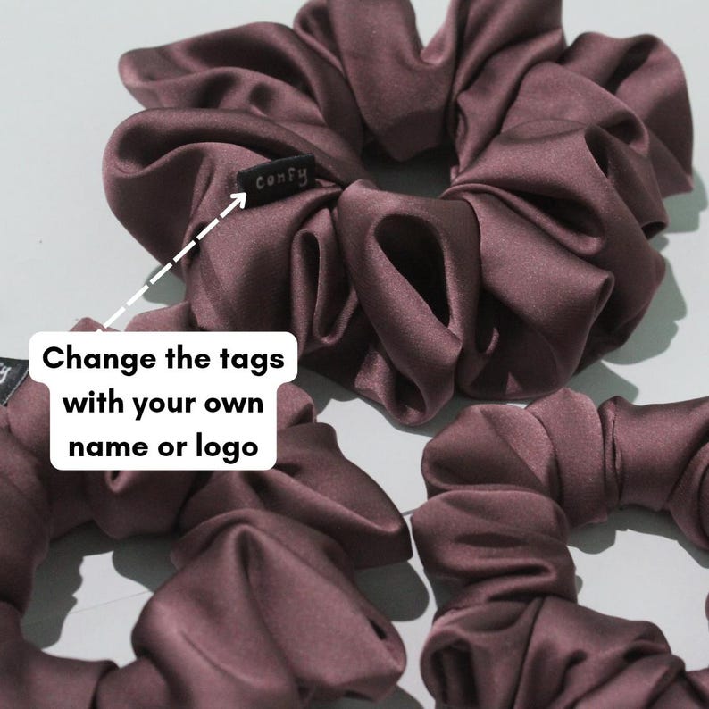 May include: Three dark plum-coloured satin hair scrunchies with gathered fabric. One scrunchie features a small black fabric tag with the white text "confy" stitched onto it. The image includes a white graphic overlay with the text "Change the tags with your own name or logo" and an arrow pointing to the tag.