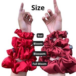 May include: A collection of red and dark red satin hair scrunchies arranged in four sizes labelled Bud, Bloom, Blossom, and Full Bloom. The scrunchies are displayed in a vertical stack on two arms, showing varying degrees of fabric fullness and volume.