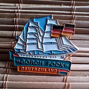 Puede incluir: Un pin de esmalte azul y blanco con un barco con una bandera alemana. El pin tiene el texto "GORCH FOCK" y "DEUTSCHLAND".