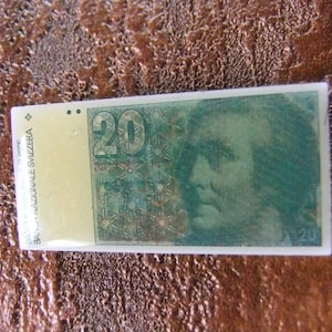 Könnte beinhalten: Eine Nahaufnahme eines 20 Schweizer Franken-Banknotens. Der Schein ist weiß mit einem grünen Porträt eines Mannes und dem Text "BANCA NAZIONALE SVIZZERA" auf der linken Seite.