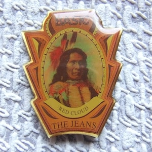 Puede incluir: Un pin de estilo vintage con un retrato de un nativo americano con un tocado de plumas. El pin tiene un borde dorado y el texto "BASFZ", "RED CLOUD" y "THE JEANS". La imagen está sobre un fondo claro.