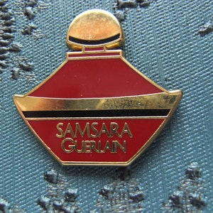 Può includere: Uno spillo a forma di flacone di profumo rosso e dorato con le parole "Samsara Guerlain" sul davanti.
