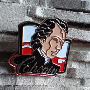 Könnte beinhalten: Eine silberne und rote Emaille-Anstecknadel mit einem Porträt des Komponisten Frédéric Chopin. Die Anstecknadel zeigt Chopin im Profil, mit langem, wallendem Haar. Die Anstecknadel trägt auch das Wort "Chopin" in einer stilisierten Schrift.