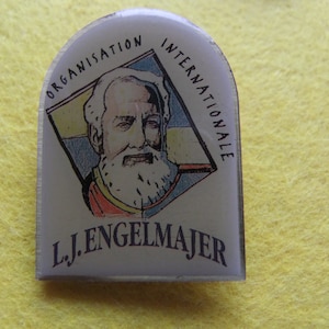 May include: A white enamel pin featuring a colourful image of a bearded man. The pin is inscribed with the text "ORGANISATION INTERNATIONALE L.J. ENGELMAJER".
