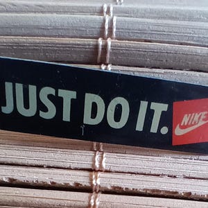 Op de afbeelding: Zwarte en witte sticker met de tekst "JUST DO IT." in witte letters en een rood Nike swoosh-logo met het woord "NIKE" in witte letters.
