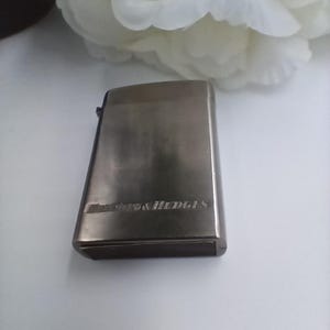 Könnte beinhalten: Ein rechteckiges, silberfarbenes Feuerzeug mit dem eingravierten Text "Benson & Hedges" auf der Oberfläche. Das Feuerzeug hat eine gebürstete Metalloberfläche und einen Klappdeckel. Im Hintergrund eine weiße Blume.