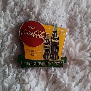 Könnte beinhalten: Vintage Coca-Cola-Anstecknadel mit einem roten kreisförmigen Logo mit der Aufschrift "Drink Coca-Cola". Die Anstecknadel hat einen gelben Hintergrund mit zwei Coca-Cola-Flaschen-Illustrationen und dem Text "Family Size" und "Regular Size". Unten steht "Two Convenient Sizes".