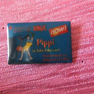 Puede incluir: Un pin de esmalte azul y rojo con una ilustración de dibujos animados de Pippi Calzaslargas haciendo una parada de manos. El pin también incluye el texto "KNAL NOW!" y "Pippi in Taka-Taka-land".
