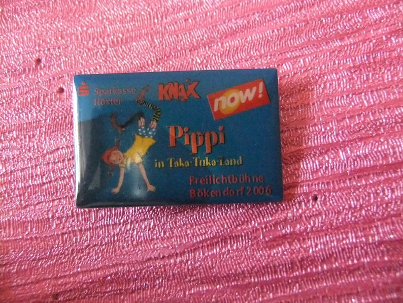 May include: A blue and red enamel pin featuring a cartoon illustration of Pippi Longstocking doing a handstand. The pin also includes the text "KNAL NOW!" and "Pippi in Taka-Taka-land".