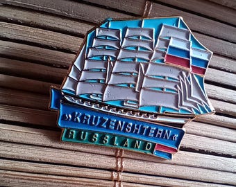 May include: A blue and white enamel pin featuring a ship with the text "KRUZENSHTERN" and "RUSSLAND" below. The ship has a white sail and a red, white, and blue flag.